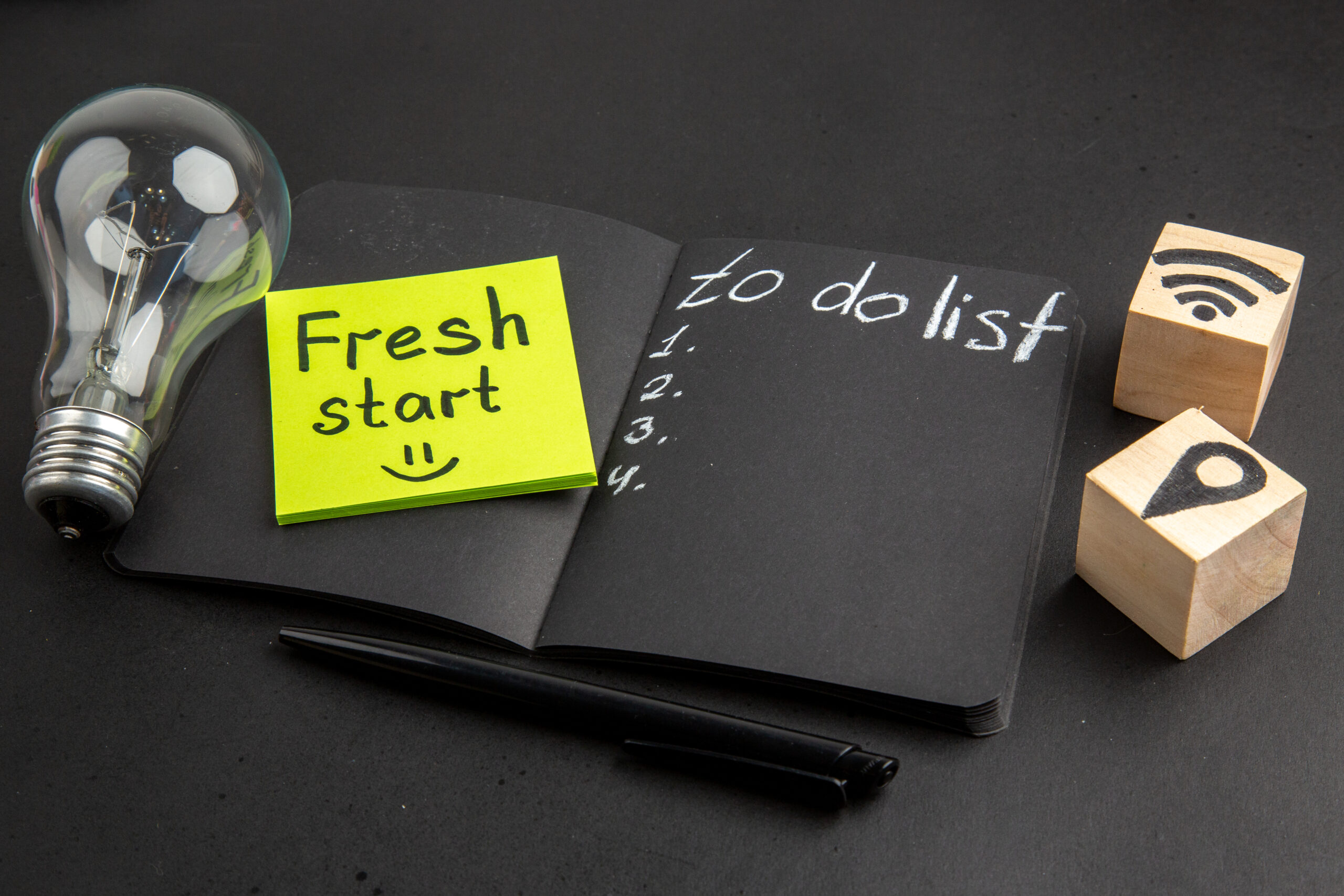 bottom-view-list-written-black-notepad-fresh-start-written-sticky-note-wifi-location-icons-wood-cubes-pen-light-bulb-black-background